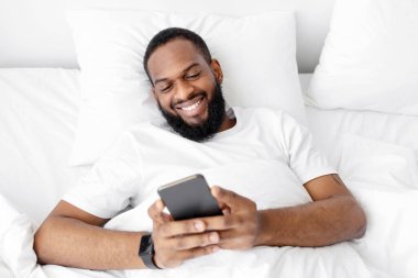Gülümseyen genç siyah sakallı adam beyaz yatakta uzanıyor, başvuru formlarını kontrol ediyor ve akıllı telefondan sosyal ağları kontrol ediyor. Covid-19 salgını, sohbet, yeni sabah haberleri ve olumlu yüz ifadesi.