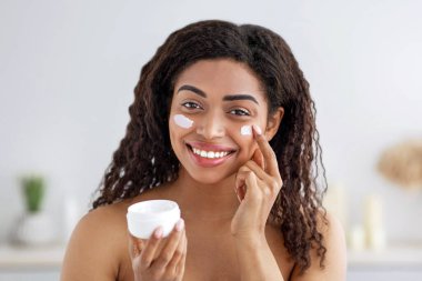 Yaşlanma önleyici bakım, kırışıklık önleyici, duştan sonra nemlendirici, güzellik koruması, covid-19. Mutlu, genç, kıvırcık, güzel, Afro-Amerikan kadın yüzüne krem sürüp yatak odasının iç kısmında kavanoz gösteriyor.