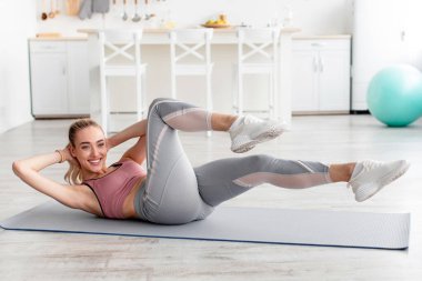 Covid-19 karantina, yeni normal, sosyal uzaklık ve evde sağlık hizmeti. Spor kıyafetli genç, sevimli sarışın kadın karın kası egzersizi yapıyor ve oturma odasının iç tarafındaki akıllı telefon web kamerasından video çekiyor.