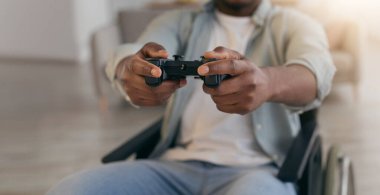 Evde boş zaman, modern cihazlar, olumlu duygular, çovid. Mutlu Afro-Amerikan adam tekerlekli sandalyede sakat, oturma odasında joystick ile bilgisayar oyunu oynuyor.