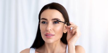 Makyaj için kozmetik ürünleri, Covid-19 tecrit sırasında evde online güzellik dersleri. Mükemmel cilde sahip genç bir bayan kirpiklerine rimel sürer ve aynaya bakar, beyaz arka planda, boşluğu kopyalar.