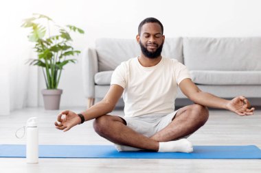 Huzurlu, sakin, bin yıllık Afro-Amerikan sakallı erkek oturma odasının iç tarafındaki yerde gözleri kapalı yoga yapıyor. Covid-19 salgını, dinlenme ve rahatlama, sağlık ve vücut bakımı, evde çalışma