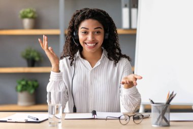 Pozitif öğretmen, öğretmen internetten uzaktan ders, eğitim, sanal webinar, covid-19 için arama yapıyor. Kulaklık takan neşeli siyah kadın oturma odasının iç tarafındaki kameraya bakıyor.