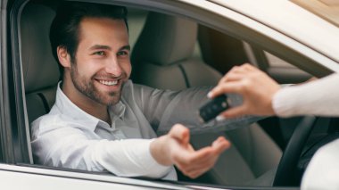 Kadın eli, bayilikte erkek eli için araba anahtarı verir. Araba endüstrisi, sigorta veya otomobil ticareti. Genç, gülümseyen, beyaz tişörtlü beyaz adam yeni arabasında oturuyor, boş bir alanda.