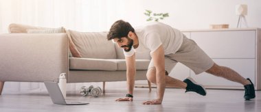 Evde spor, online ders ve kendini tecrit sırasında vücut bakımı. Ciddi Arap sakallı genç adam bacak egzersizi yapıyor. Dizüstü bilgisayarına bakıyor. Oturma odasında yerde bir şişe su var.