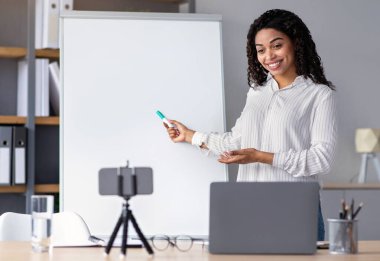 Modern teknoloji ile çevrimiçi ders, ders çalışma, sosyal mesafe ve yeni normal. Dost canlısı gülümseyen Afrikalı Amerikalı kadın tahtayı işaret ediyor ve web kamerasında öğrenciler için klip çekiyor.