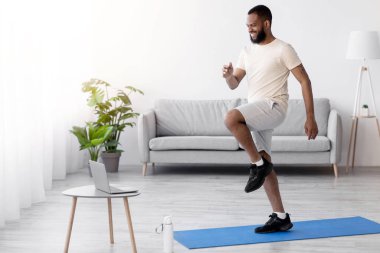 Beyaz elbiseli, gülümseyen genç siyah atlet bacak egzersizleri, ısınma egzersizleri yapıyor, içerideki odada bilgisayardan video seyrediyor. Vücut bakımı, fitness günlüğü, sosyal mesafe, covid-19 tecrit sırasında evde çalışma