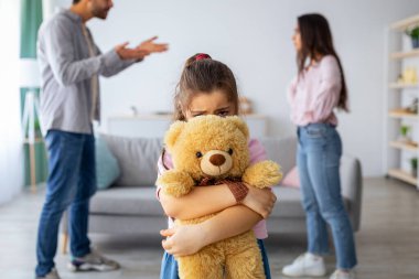 Çocuk ve aile sorunları kavramı. Oyuncak ayının arkasına saklanan küçük bir kız, ebeveynlerin kavgalarından muzdarip, yalnız ve depresif, seçici bir şekilde çocuğa odaklanmış.