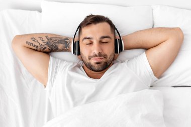 Huzurlu, sakin, kulaklıkları kapalı genç Avrupalı kaslı adam beyaz yatakta uzanıyor, yatak odasında uyuyor, en sevdiği müziği dinliyor ve boş zamanların tadını çıkarıyor. Sağlıklı uyu, dinlen ve evinde dinlen.