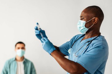 Covid-19 'u engelle, aşı merkezinde salgından aşı olma. Olgun Afro-Amerikan erkek hemşire ya da maskeli doktor, klinikteki genç hastaya aşı enjekte etmek için şırıngaya aşı çeviriyor.