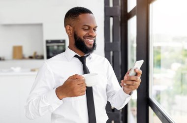 Portrait of smiling African American man using mobile phone holding cup of coffee standing by window, watching video chatting online, checking email enjoying weekend, modern home or coworking interior
