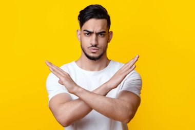 Ciddi Arap adam dursun ya da durmasın sarı stüdyo arka planı, kopyalama alanı. Orta Doğulu, beyaz tişörtlü bir gencin elleri kesişiyor, bir şeyi sevmiyor ya da inkar ediyor.