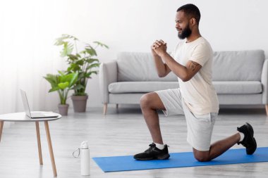 Gülümseyen bin yıllık Afro-Amerikan sakallı adam paspasın üstünde bacak egzersizi yapıyor, odanın içinde bir şişe suyla, dizüstü bilgisayarda egzersiz yapıyor. Çevrimiçi sınıf, evde yeni normal ve vücut bakımı