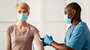 Aşı, aşı, hastalık önleme. Maskeli yetişkin kadın kovid 19 ya da grip aşısı oluyor. Profesyonel Afro-Amerikan üniformalı doktor klinikteki hastaya antiviral iğne yapıyor.
