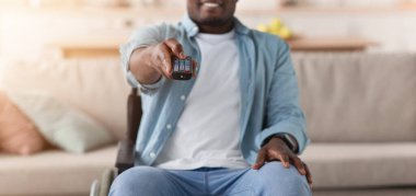 Evde eğlence, TV şovu ve boş zamanlarında film, dinlenme ve rahatlama, covid. Neşeli yetişkin siyahi adam tekerlekli sandalyede engelli TV 'yi uzaktan kumandalı oturma odasının içinde, seçici odaklı