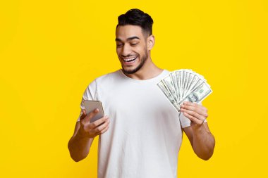 Beyaz tişörtlü şanslı genç adam internette hisse senedi ve pazar alışverişi yapıyor, cep telefonu kullanıyor, bir sürü para tutuyor ve gülümsüyor, sarı stüdyo geçmişi var, kopyalama alanı kullanıyor.