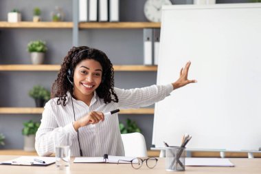 Neşeli genç bayan öğretmen tahtaya işaret ediyor, gramerini açıklıyor, ev ofisinden çevrimiçi İngilizce dersi veriyor. Kulaklıklı Afrikalı Amerikalı kadın webcam 'e bakıyor, içeride blog için çekim yapıyor.