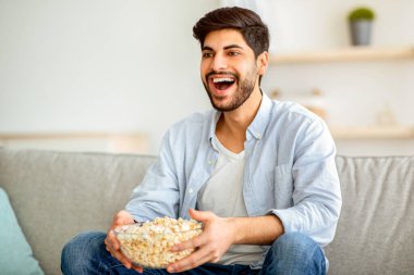 Neşeli Arap adam TV 'de en sevdiği filmi izliyor, patlamış mısır yiyor, oturma odasında kanepede dinleniyor. Mutlu doğu çocuğu rahat bir kanepede dinleniyor, hafta sonunun keyfini çıkarıyor, ev işleri yapıyor.