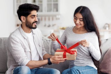 Sevgililer günü, doğum günü, yıldönümü hediyesi. Gülümseyen bin yıllık sakallı Arap koca, güzel bir bayana hediye veriyor. Oturma odasının içinde kırmızı kurdele bağlayan kadın.