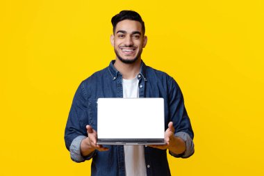 Neşeli Arap adam, sarı arkaplanda boş ekranlı yepyeni bir laptop gösteriyor. Orta Doğulu genç adam, bilgisayarlar için yeni bir yazılım öneriyor.