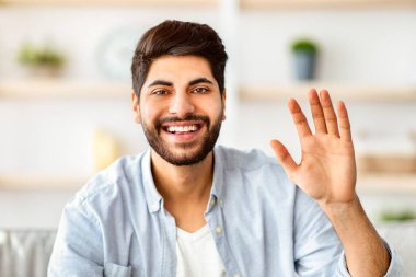 Selamlama hareketi konsepti. Mutlu Arap adam el sallıyor ve kameraya bakıyor, evdeki kanepede oturuyor. Adam merhaba deyip gülüyor. Video çağrısı ve modern uzaktan iletişim.