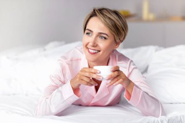 İyi sabahlar. Rahatlamış gülümseyen kadın portresi beyaz çarşaflarla rahat bir yatakta uzanıyor, başka yere bakıyor ve kahve içiyor, elinde bir fincan sıcak çay tutuyor, boş zamanın tadını çıkarıyor, bedava fotokopi çekiyor.