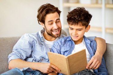 Evde faydalı bir zaman. Baba oğul kitap okuyor ve gülümsüyor, oturma odasındaki rahat kanepede dinleniyorlar. Ebeveynlik ve aile ilişkisi, hafta sonunu birlikte geçirmek.