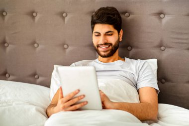 Hafta sonu eğlence konsepti. Rahat Arap adam dijital tablet kullanıyor, yatakta oturuyor ve evdeki yatak odasında internette sörf yapıyor. Gadget, teknolojiler ve modern yaşam tarzı.