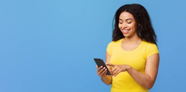 Güzel uygulama. Mutlu Afro-Amerikalı kadın akıllı telefon kullanıyor, mesajlaşıyor ya da sosyal ağları kaydırıyor, mavi stüdyo arka planında duruyor.