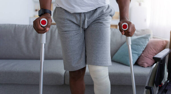 Bone fracture, injury, recovery and treatment at home after incident. Mature black man with broken leg with plaster of cast walk with crutches in living room interior with wheelchair, cropped