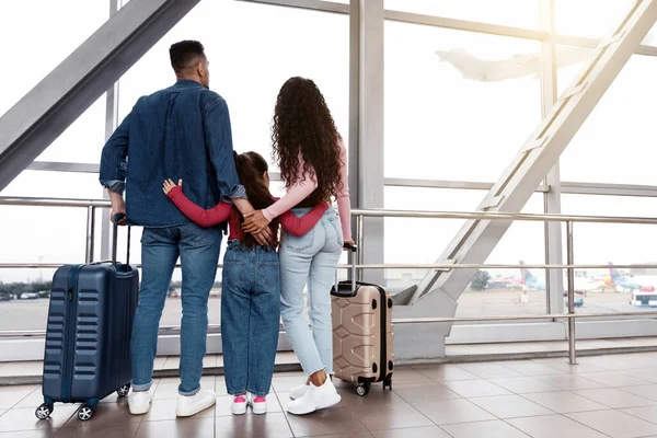 Havalimanı Terminali 'ndeki pencereden bakan kızıyla aile manzarası, ebeveynler ve onların küçük bayan çocukları tatil için hazırlar, bavullarıyla dururlar ve kucaklaşırlar, boşluğu doldururlar.