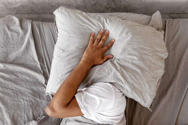 Closeup of irritated young black man lying in bed and covering head and ears with pillow, hearing and suffering from too loud sound. Angry millennial guy cant sleep, tired of noisy neighbors
