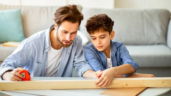 Yoğunlaşmış baba ve oğul tahta parçasını bantla ölçüyor, çocuk ev işi öğretiyor. Aile, evdeki ahşap detaylardan kuş evi yapıyor.