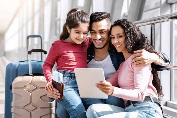 Havalimanında dijital tabletle rahatlayan üç kişilik mutlu Arap ailesi, genç ortadoğulu ebeveynler ve modern cihaz kullanan sevimli bayan çocuk terminalde uçuşu beklerken, yakın çekim