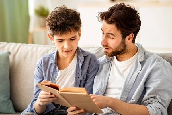 Aile eğlencesi. Genç baba ve genç çocuk birlikte kitap okuyor, oturma odasındaki rahat kanepede dinleniyorlar. Ebeveynlik ve aile ilişkisi, tek ebeveyn kavramı