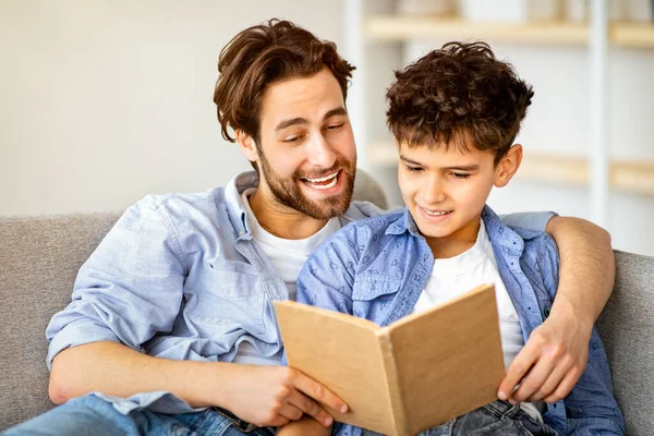 Evde faydalı bir zaman. Baba oğul kitap okuyor ve gülümsüyor, oturma odasındaki rahat kanepede dinleniyorlar. Ebeveynlik ve aile ilişkisi, hafta sonunu birlikte geçirmek.