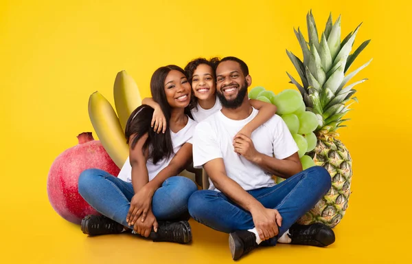 Üç neşeli aile, çeşitli taze meyvelerle çevrili canlı sarı bir arka plana karşı birlikte otururlar. Sağlıklı beslenmenin ve beslenmenin faydalarını somutlaştırıyorlar..
