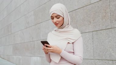 Başörtüsü takan genç bir kadın sokakta akıllı telefonuyla meşgul. Pürüzsüz taş bir duvara karşı duruyor. Canlı bir şehirde rahat ve çağdaş bir yaşam tarzı sergiliyor..