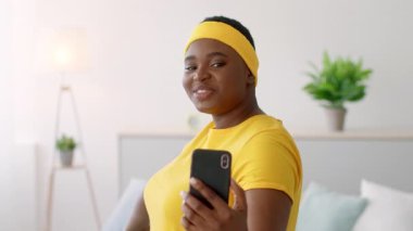 Büyük boy Afro-Amerikan bir kadın, elinde telefon tutarken pozitif tavrını sergileyerek evde antrenman yapıyor. Canlı bir fitness kıyafetiyle spor ve vücut pozitifliğini teşvik ediyor..