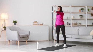 Pembe spor kıyafetli ve siyah taytlı genç bir kadın evde egzersiz yapıyor. Ağırlık kaldırırken gülümsüyor. Çevresi parlak ve ferah, sağlıklı bir yaşam tarzını destekliyor..