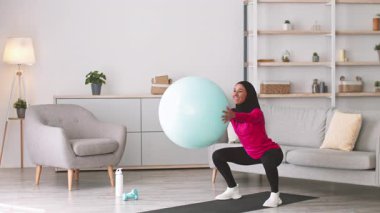 Oturma odasında tesettürlü genç bir kadın çömelirken denge topunu kullanıyor. Konforlu evinde sağlıklı bir yaşam tarzını benimsiyor. Formda ve kararlı olduğunu gösteriyor..