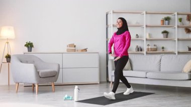 Pembe eşofmanlı genç bir Müslüman kadın evde egzersiz yapıyor, tesettüre girerek sağlıklı bir yaşam tarzını geliştiriyor. Modern iç mekan onun motive edici çevresine katkı sağlıyor..
