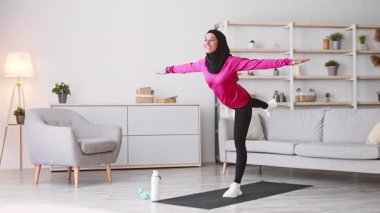 Başörtüsü takan genç bir kadın evde egzersiz yapıyor. Kollarını uzatırken tek bacağı üzerinde dengede duruyor. Parlak ve davetkar alan egzersiz yoluyla sağlıklı bir yaşam tarzını teşvik eder..