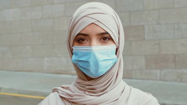 Açık renk tesettürlü ve maskeli genç bir kadın şehir caddesinde güven içinde duruyor. Canlı bir şehir ortamında açık havanın tadını çıkarırken stilini ve zarafetini sergiliyor..