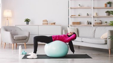Orta Doğulu genç bir kadın, şık oturma odasında dengeli bir top üzerinde antrenman yapıyor. Huzurlu bir ortamda egzersiz yaparken sağlıklı bir yaşam tarzını teşvik ediyor..