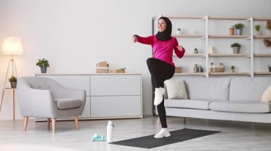 Genç, tesettürlü bir Orta Doğulu kadın evde enerjik bir egzersiz yapıyor. Yoga minderinin üzerinde egzersiz yapıyor, rahat bir oturma odasında sağlıklı bir yaşam tarzını teşvik ediyor..