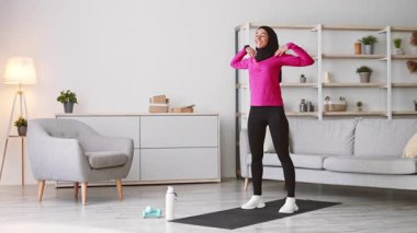 Genç, tesettürlü bir Orta Doğulu kadın şık evinde egzersiz yapıyor. Esnerken gülümsüyor, modern bir çevrede sağlıklı bir yaşam tarzını teşvik ediyor..