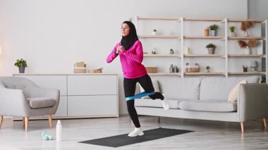 Genç bir Ortadoğulu kadın oturma odasında tesettür egzersizi yapıyor. Parlak bir bluz giyiyor ve egzersiz yapmak için direnç bantlarını kullanarak vücut geliştirmeye çalışıyor..