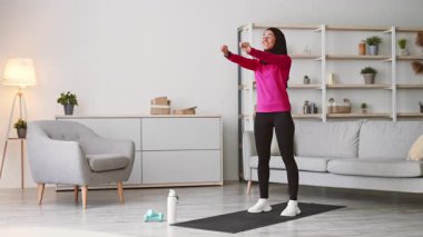 Parlak bir oturma odasında, spor kıyafeti giymiş genç bir Orta Doğulu kadın egzersiz yapıyor. Etrafı modern mobilyalarla çevrili, tesettür takarken fitness rutinine odaklanıyor..