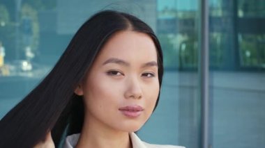 Odaklanmış Asyalı bir kadın, profesyonel giyinmiş, kendine güvenen bir şekilde bir şirket ortamında duruyor. Gündüz saatlerinde modern ofis ortamının tadını çıkarırken işine daldı..